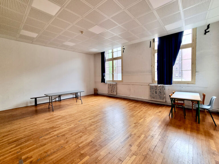 Salle de résidence du Labo Victor Hugo avec parquet, grandes fenêtres encadrées de rideaux bleus et mobilier minimal comprenant une table et quelques chaises.